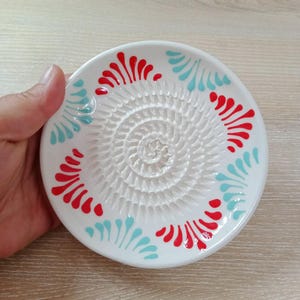 Może przedstawiać: Biała ceramiczna tarka do czosnku z spiralną powierzchnią do ścierania. Talerz jest ozdobiony czerwonymi i turkusowymi kwiatowymi wzorami wokół krawędzi. Tarka jest trzymana w dłoni.