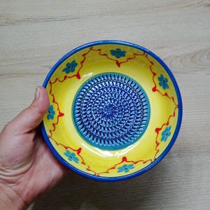 Może przedstawiać: Ręcznie malowana ceramiczna płyta do tarcia. Płyta ma żółte tło z niebieskimi kwiatowymi akcentami i niebieską obwódką. Środek to teksturowana niebieska powierzchnia do tarcia.
