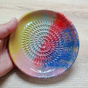 Może przedstawiać: Kolorowa ceramiczna tarka do czosnku z spiralną powierzchnią do ścierania. Płytka ma żywą mieszankę kolorów, w tym żółty, różowy, czerwony i niebieski, z efektem nakrapianej farby.