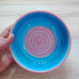 Może przedstawiać: Ceramiczna miska do tarcia o żywym designie. Miska ma jasnoniebieskie wnętrze i różowy brzeg. Środek miski ma teksturowaną, spiralnie wzorzystą tarkę w delikatnym różowym kolorze. Miska jest przeznaczona do użytku w kuchni.