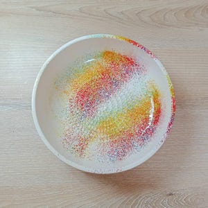 Może przedstawiać: Biała ceramiczna miska z kolorowym, nakrapianym wzorem. Wnętrze miski ma żywy wzór czerwonych, żółtych, zielonych i niebieskich plamek, tworząc efekt tęczy. Misa stoi na jasnej drewnianej powierzchni.