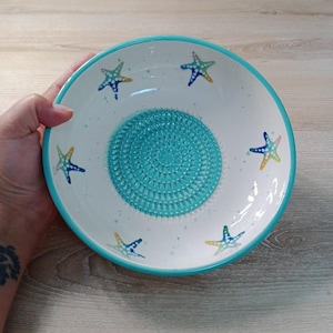 Może przedstawiać: Biała ceramiczna miska z turkusową krawędzią i teksturowanym turkusowym środkiem. Misa jest ozdobiona kolorowymi rozgwiazdami i małymi niebieskimi plamkami. Element dekoracyjny do kuchni lub jadalni.
