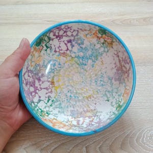Może przedstawiać: Kolorowa ceramiczna miska o teksturowanym wnętrzu i jasnoniebieskim obramowaniu. Wnętrze miski ma nakrapiany wzór w odcieniach fioletu, żółci, zieleni i pomarańczy. Miskę trzyma ręka, prezentując jej rozmiar i design.