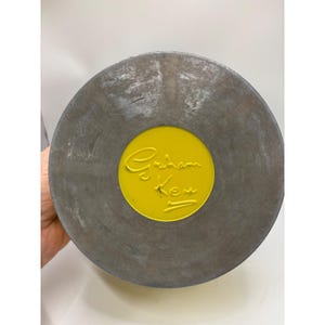 May include: A round, gray metal object with a yellow center. The yellow center features the embossed text "Graham Kerr" in a cursive script. The object has a worn, textured appearance.