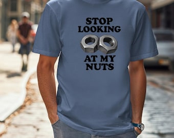 May include: A blue t-shirt with the text "STOP LOOKING AT MY NUTS" in black, along with an image of two metallic nuts. The shirt is worn by a person in a street setting.