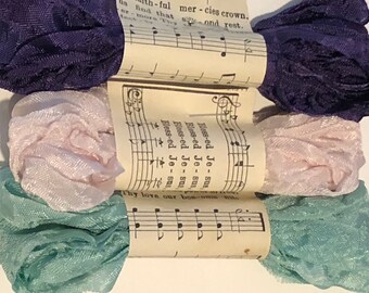 May include: Three spools of ribbon, one dark purple, one light pink, and one light teal, wrapped around pages of sheet music with the words "Blessed Jesus" printed on them.