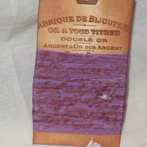 Peut inclure: Un ruban violet avec une petite perle blanche sur une étiquette brune avec le texte "Fabrique de Bijouterie Or a tous titres Doublé Or Argent & Or sur Argent".