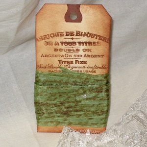 May include: A brown paper tag with the text "ABRIQUE DE BIJOUTERIE OR A TOUS TITRES DOUBLE OR ARGENT & OR SUR ARGENT TITRE FIXE Seul Double Or garanti inalterable RACHEE'S USAGE" and a green ribbon wrapped around it.