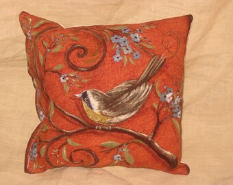 Może przedstawiać: Dekoracyjna poduszka z pomarańczowym tłem, na której znajduje się brązowo-żółty ptak siedzący na gałęzi z zielonymi liśćmi i niebieskimi kwiatami.