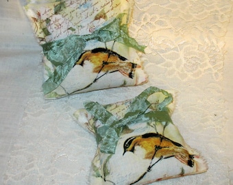 May include: Two small fabric sachets with a floral and bird print. Each sachet has a green ribbon tied around it. The sachets are on a white lace background.
