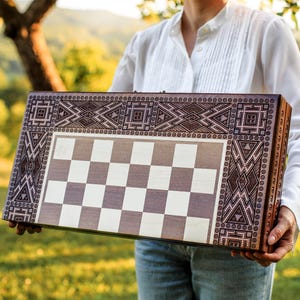 Puede incluir: Un tablero de ajedrez y backgammon de madera con intrincados diseños geométricos en los bordes. El tablero presenta un patrón de cuadros blancos y grises. El tablero se sostiene frente a un fondo verde borroso.