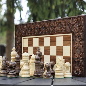Puede incluir: Un juego de ajedrez de madera con bordes de color marrón oscuro tallados intrincadamente y un tablero a cuadros. Las piezas de ajedrez son una mezcla de marrón claro y oscuro, dispuestas sobre una superficie oscura.