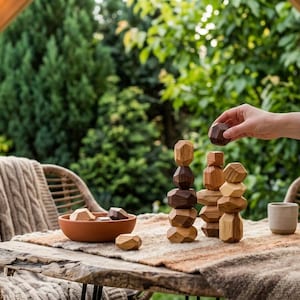 May include: Wooden stacking toys in various shades of brown and tan are arranged on a rustic wooden table. A hand is placing a piece on top of a stack. A terracotta bowl and a small ceramic cup are also on the table.