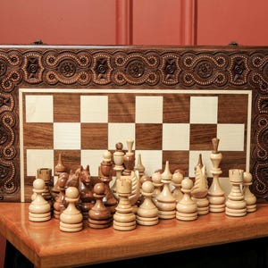 May include: A wooden chess set with intricately carved dark brown borders and a chessboard with alternating light and dark squares. The chess pieces are arranged on the board, ready for play. The set is displayed on a wooden table.