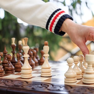 Puede incluir: Un juego de ajedrez con piezas de madera claras y oscuras sobre un tablero a cuadros. El tablero está sobre una superficie de madera oscura tallada. Una mano, con una manga blanca con rayas azul marino, rojas y blancas, mueve una pieza.