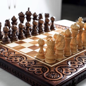 May include: A wooden chess set featuring a board with intricate carved borders and two sets of pieces in dark brown and light tan. The pieces display detailed textures and patterns, arranged on a square grid of alternating light and dark wood squares.