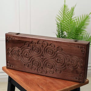 May include: A rectangular, wooden backgammon set with intricate, carved floral and geometric patterns. The dark brown set is displayed on a wooden table, with a green plant in the background. The set is closed and ready for travel.