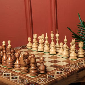 May include: A wooden chess set with light and dark brown pieces arranged on a checkered board. The board is set on a wooden table with decorative inlaid patterns. The background is a solid red-brown colour.