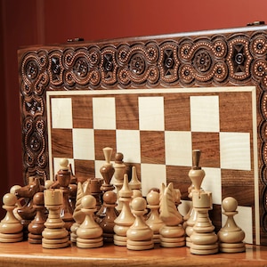 Puede incluir: Un juego de ajedrez de madera con detalles tallados. El tablero presenta un patrón de cuadrados claros y oscuros. Las piezas de ajedrez están dispuestas frente al tablero. El conjunto se exhibe sobre una superficie de madera.