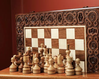 May include: A wooden chess set with intricately carved detailing. The chessboard features a light and dark square pattern. Chess pieces are arranged in front of the board. The set is displayed on a wooden surface.