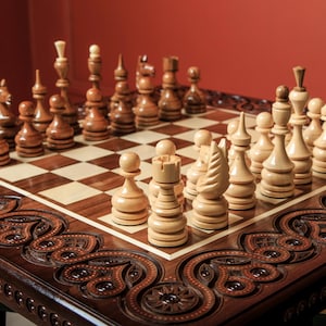 Puede incluir: Un juego de ajedrez de madera con piezas talladas dispuestas sobre un tablero a cuadros. El tablero está sobre una mesa marrón oscuro con tallas decorativas. Las piezas de ajedrez son de color marrón claro y oscuro.