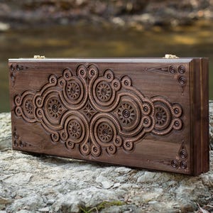 May include: A dark brown wooden backgammon set with intricate, carved floral and geometric designs. The set is rectangular, with a detailed pattern across the front. The set is resting on a rock, with a blurred natural background.