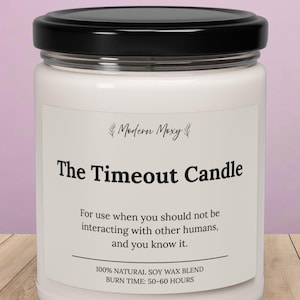 May include: A white candle in a glass jar with a black lid. The label reads "The Timeout Candle" with text below. The candle is on a wooden surface with a light purple background.