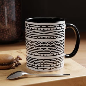 May include: White coffee mug with a black interior and handle. The mug features a repeating black geometric pattern. A spoon, a macaron, and coffee beans are on a wooden surface next to the mug.