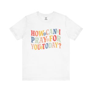 Könnte beinhalten: Weißes T-Shirt mit dem Text "HOW CAN I PRAY FOR YOU TODAY?" in farbenfrohen Buchstaben im Retro-Stil. Das Design enthält Blumenakzente. Das Shirt ist aus einem weichen Material.