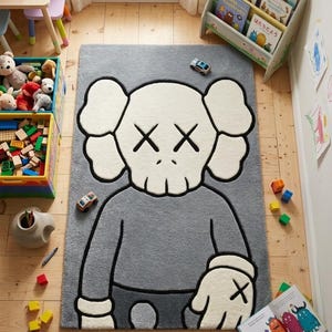 May include: Gray rectangular rug featuring a cartoon character design with white and black accents. The rug is placed on a wooden floor in a child's room, surrounded by toys and a small bookshelf.