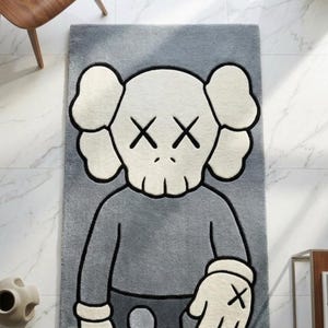 May include: Gray rectangular rug featuring a cartoon character with crossed-out eyes and a skull-like face. The rug has a gray body with white and black accents. The character's arms and hands are visible, with a black 'X' on the hand.
