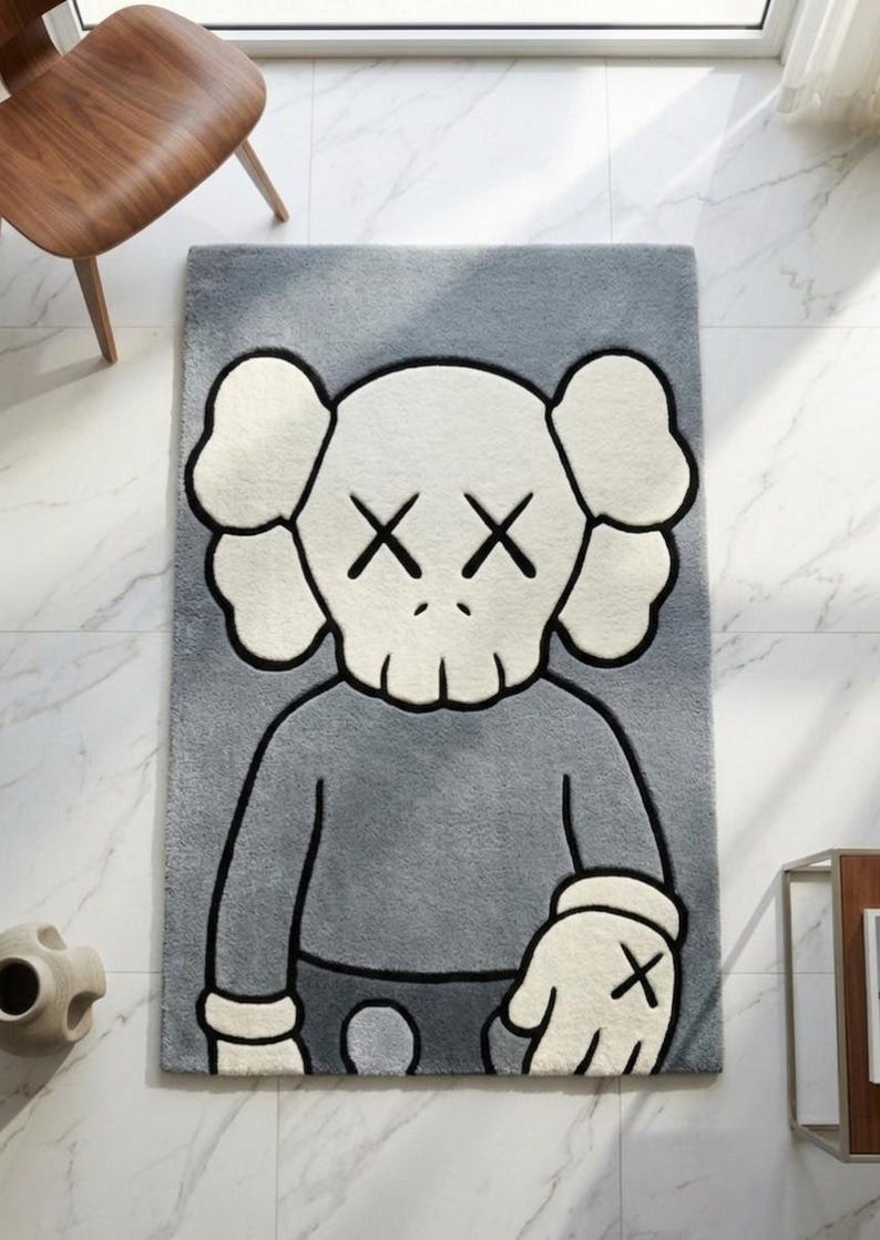 May include: Gray rectangular rug featuring a cartoon character with crossed-out eyes and a skull-like face. The rug has a gray body with white and black accents. The character's arms and hands are visible, with a black 'X' on the hand.