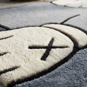 May include: Close-up of a gray and white rug with a cartoon-style design. The rug features a white shape with a black cross and black outline, set against a gray background. The rug has a soft, textured appearance.