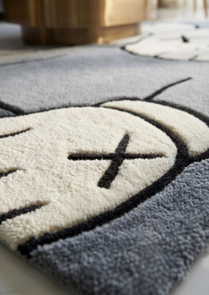 May include: Close-up of a gray and white rug with a cartoon-style design. The rug features a white shape with a black cross and black outline, set against a gray background. The rug has a soft, textured appearance.