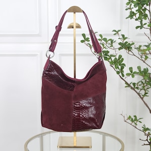 Könnte beinhalten: Eine tiefburgunderrote Hobo-Tasche mit einer Kombination aus glatten Wildledereinsätzen und glänzenden, strukturierten Lederabschnitten in Schlangenlederoptik. Die Tasche hat einen verstellbaren Schulterriemen mit silberfarbenen Metallringen.