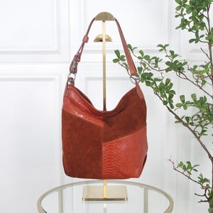 Könnte beinhalten: Eine Hobo-Tasche aus einer Kombination von rostfarbenem Wildleder und orangefarbenem, geprägtem Schlangenleder. Die Tasche verfügt über einen verstellbaren Schulterriemen mit silberfarbenen Metallringen und eine lockere, formlose Silhouette.