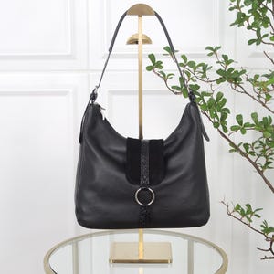 Könnte beinhalten: Eine schwarze Hobo-Tasche aus genarbtem Leder mit einer zentralen vertikalen Klappe in Wildleder- und Schlangenlederoptik. Die Tasche verfügt über einen silberfarbenen Metallringverschluss, einen einzelnen Schulterriemen und silberne Beschläge.