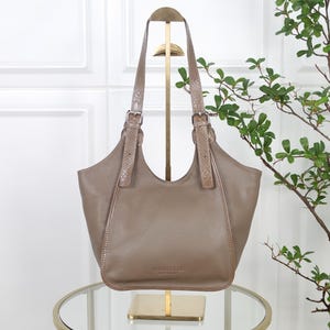Könnte beinhalten: Eine taupefarbene Ledertasche mit genarbter Textur, ausgestattet mit zwei langen Schulterriemen und verstellbaren silberfarbenen Metallschnallen. Die Tasche hat eine geschwungene Öffnung und eine Vordertasche mit der Prägung VERA PELLE MADE IN ITALY.