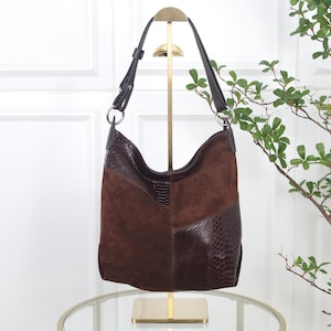 Könnte beinhalten: Eine dunkelbraune Hobo-Tasche aus einer Kombination von glatten Wildleder-Einsätzen und strukturiertem, schlangenledergeprägtem Leder. Die Tasche verfügt über einen verstellbaren Schulterriemen mit silberfarbenen Metallringen.