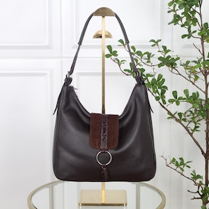 Könnte beinhalten: Eine dunkelbraune Hobo-Tasche aus genarbtem Leder mit einem einzelnen Schulterriemen und einem zentralen Klappverschluss. Die Klappe ist mit einem dunkelbraunen Wildledereinsatz, einer schlangenhautartigen Verzierung und einem silbernen Metallring versehen.