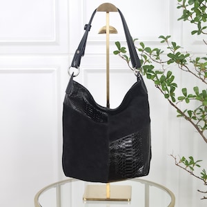 Könnte beinhalten: Eine schwarze Hobo-Tasche aus einer Kombination von mattem Wildleder und glänzenden, schlangenledergeprägten Einsätzen. Die Tasche verfügt über einen verstellbaren Schulterriemen mit silberfarbenen Metallringen und eine weiche Form.