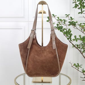 Könnte beinhalten: Eine braune Wildledertasche mit einer breiten, geschwungenen Öffnung und zwei verstellbaren Schulterriemen mit strukturierter Reptilienoptik. Die Tasche verfügt über silberfarbene Metallschnallen an den Riemen und ein dezentes, eingeprägtes Logo auf der Vorderseite.