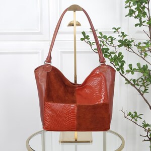 Könnte beinhalten: Eine Schultertasche im Hobo-Stil mit einem Patchwork-Design aus rostfarbenem Leder und Wildleder. Die Tasche zeigt abwechselnde Paneele aus glattem Leder mit einer schlangenartigen Prägung und weichem, mattem Wildleder, ergänzt durch zwei Trageriemen.