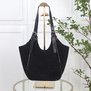 Può includere: Una borsa tote in camoscio nero con silhouette curva, due tracolle con fibbie color argento e bordi neri testurizzati lungo i lati. La borsa presenta una finitura opaca e liscia sul corpo principale e un motivo sottile sui manici.