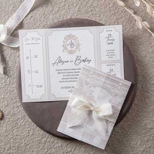 Puede incluir: Elegante juego de invitaciones de boda con una tarjeta tríptica con un escudo floral y los nombres "Aleyna ve Berkay". Incluye una tarjeta más pequeña atada con una cinta y la fecha "22 2028".
