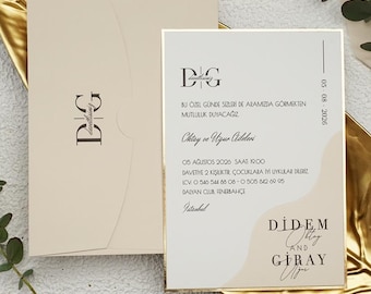May include: Elegant wedding invitation suite featuring a cream-colored invitation card with gold trim and a matching envelope. The invitation includes stylized text and a monogram, presented on a gold leaf-shaped tray with greenery.