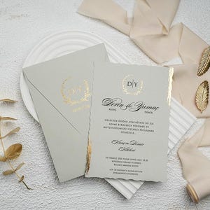 Puede incluir: Un conjunto de invitaciones de boda gris claro con detalles dorados. El conjunto incluye una tarjeta de invitación, un sobre y elementos decorativos como detalles de hojas doradas y cinta color crema. La invitación tiene texto en negro.
