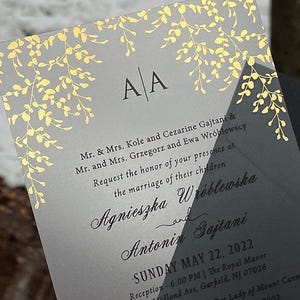 May include: A wedding invitation with a gray background and gold floral accents. The invitation includes the initials "A A" at the top and details about the wedding ceremony and reception, including the date, time, and location.