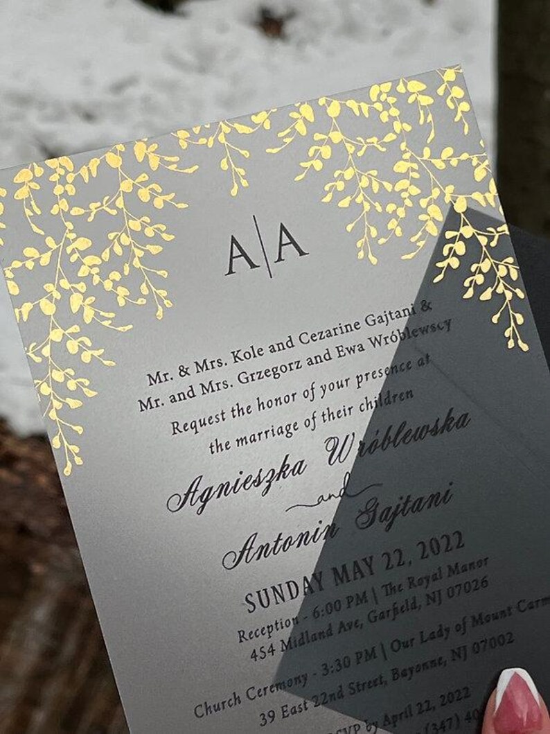 May include: A wedding invitation with a gray background and gold floral accents. The invitation includes the initials "A A" at the top and details about the wedding ceremony and reception, including the date, time, and location.