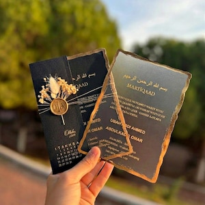 Puede incluir: Dos tarjetas de invitación de boda rectangulares con fondo negro y letras doradas. Una tarjeta presenta un calendario, un sello de cera dorado con iniciales y flores secas. La otra tarjeta tiene una superposición translúcida con bordes dorados.